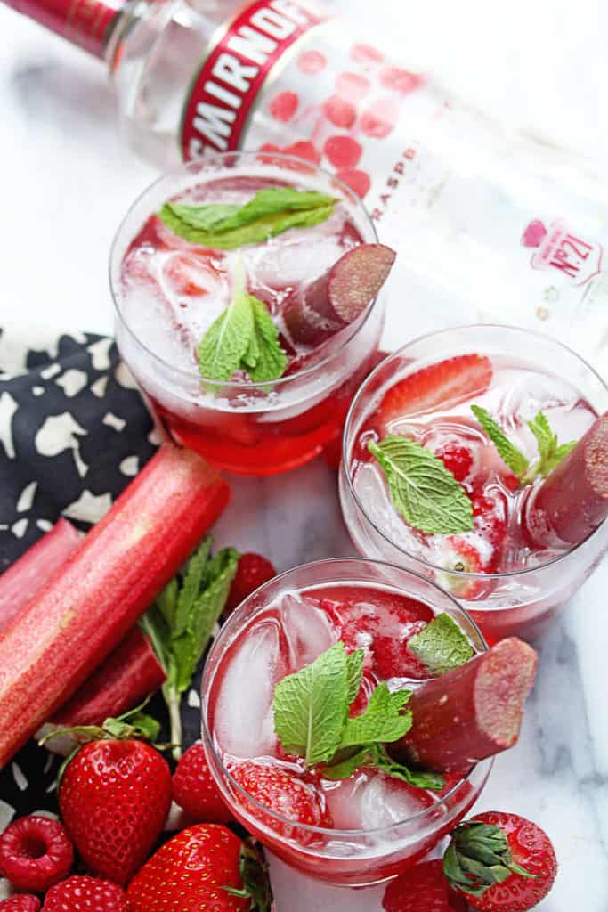 Overhead shot of three glasses of Berry Rhubarb Punch garnished with pieces of mint and rhubarb and a bottle of smirnoff vodka, fresh strawberries and rhubarb nearby