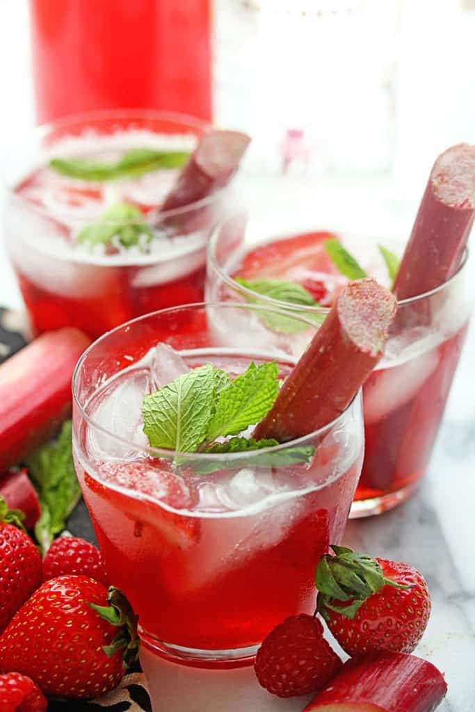 Three glasses of Berry Rhubarb Punch garnished with pieces of mint and rhubarb and fresh strawberries and rhubarb nearby