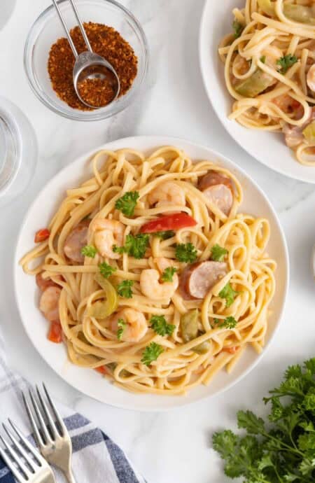 Overhead view of a plate of cajun shrimp pasta served with parsley garnish and surrounded by a bowl with cajun seasoning, more pasta, and silverware