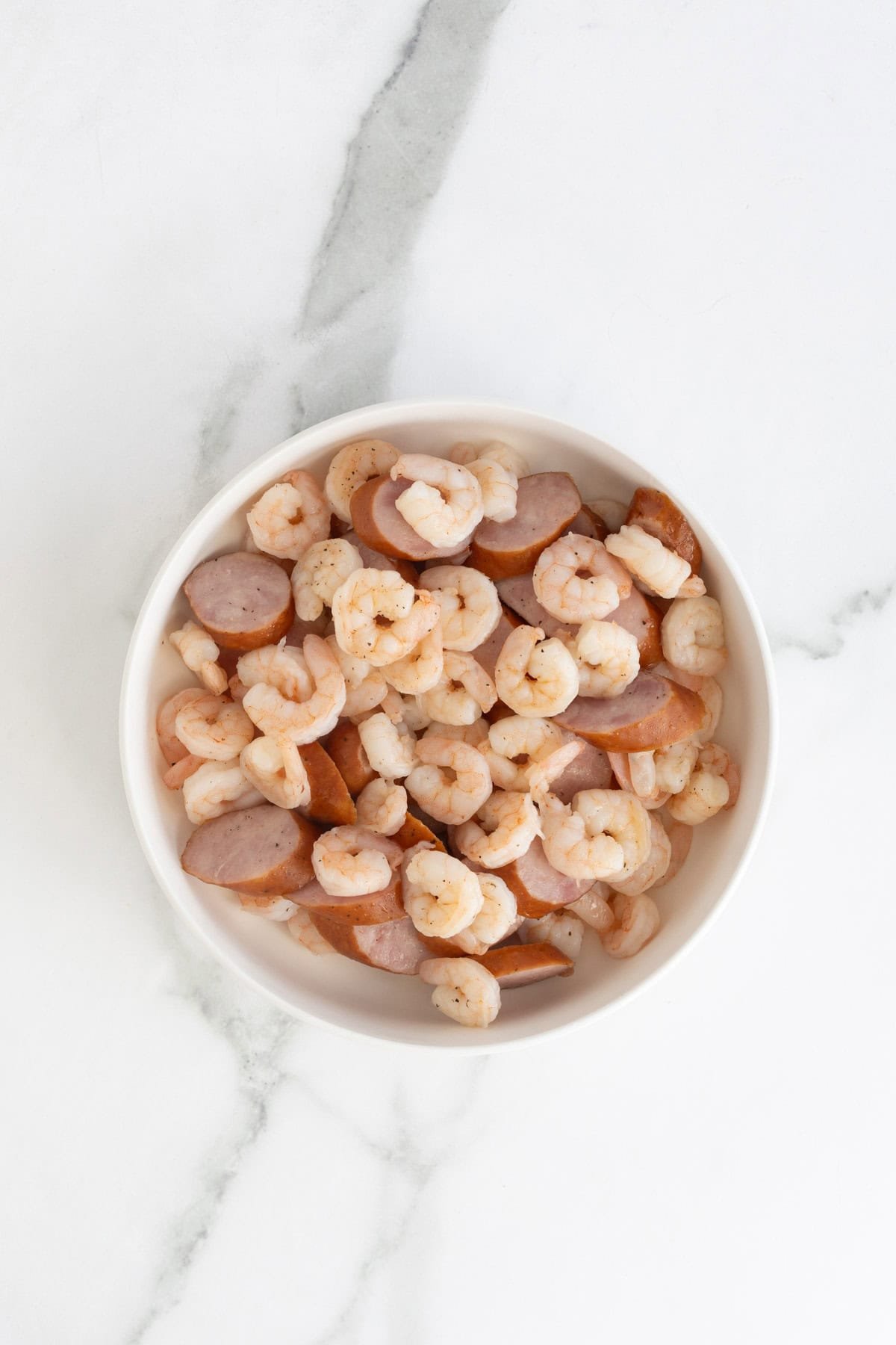 Cooked shrimp and sausage pieces in a white bowl on a marble surface