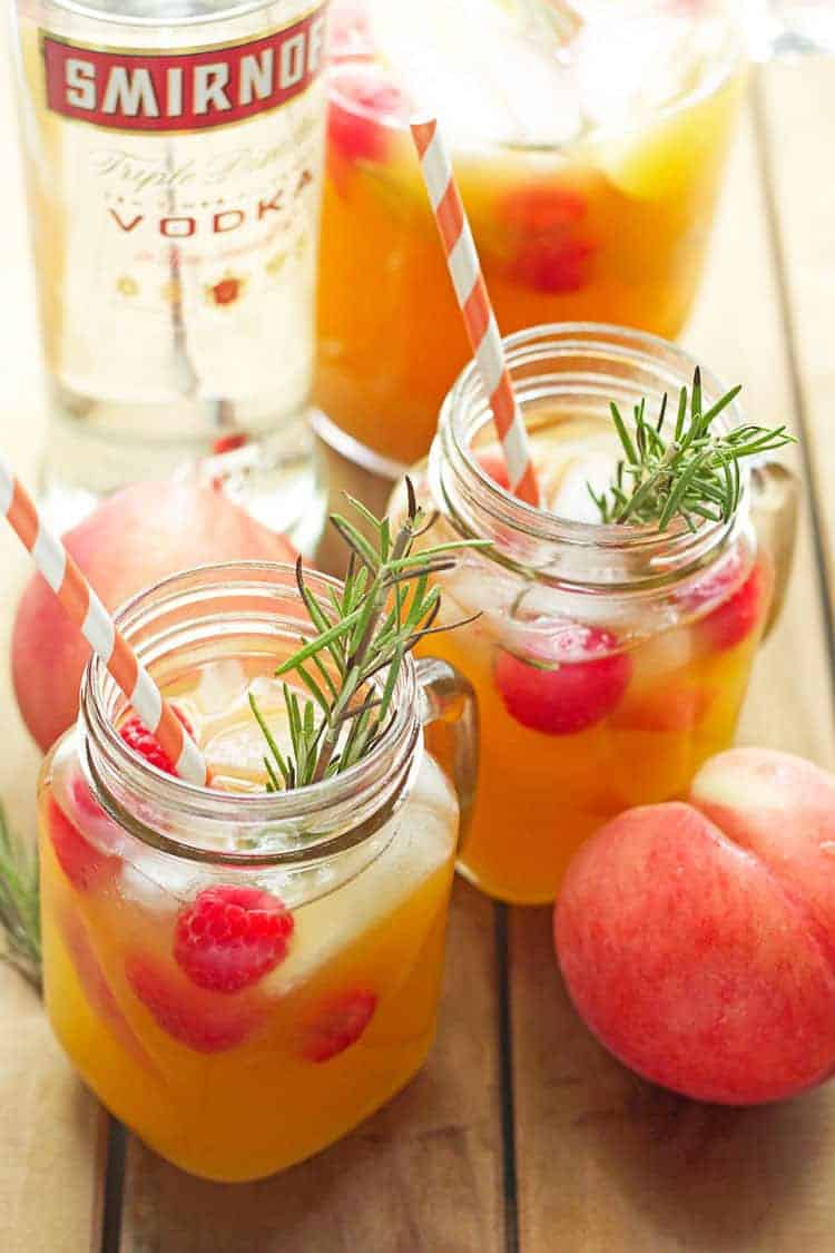 Two glass mugs filled with Peach Punch garnished with fresh raspberries and striped straws and a pitcher of the punch and a bottle of vodka in the background