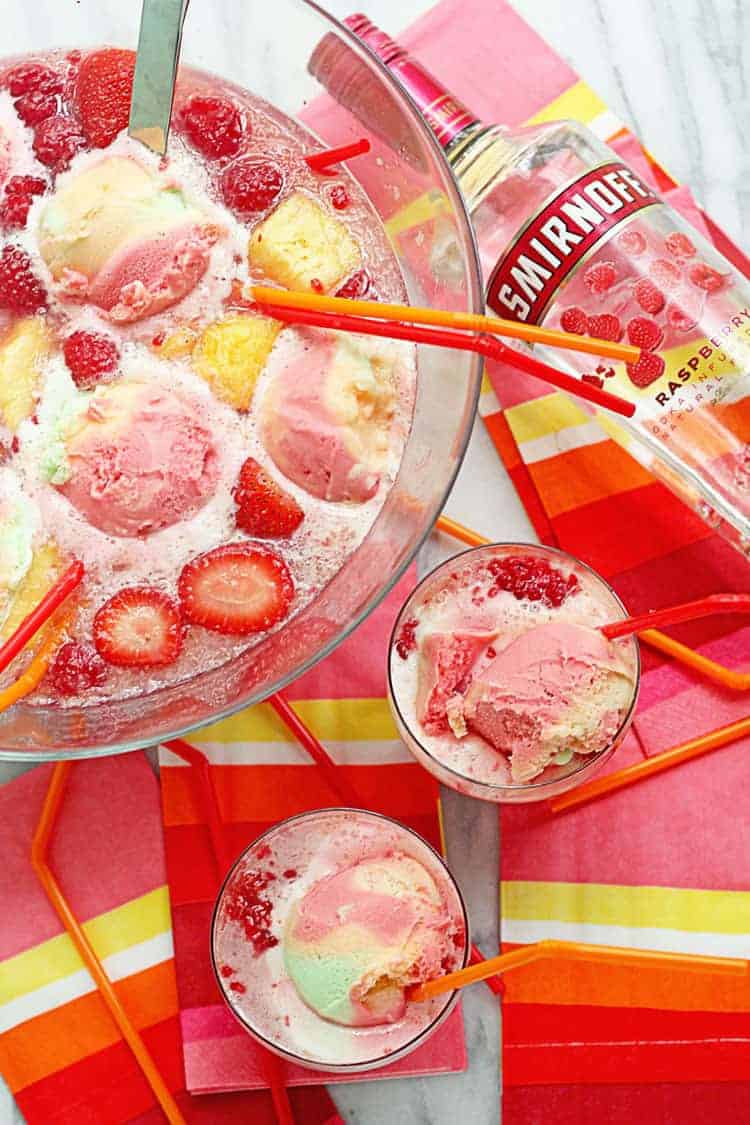 Rainbow sherbet punch contained in a large glass punch bowl and two small glasses with straws and a bottle of smirnoff raspberry vodka next to them