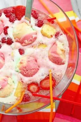 Overhead shot of Spiked Sherbet Frappe Punch in a large glass punch bowl with scoops of rainbow sherbet, fresh berries and straws in it