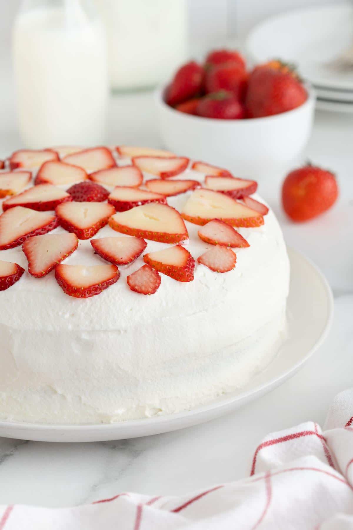 A strawberry shortcake cheesecake topped with fresh sliced strawberries arranged in a decorative pattern, with a bowl of strawberries and a bottle of milk in the background