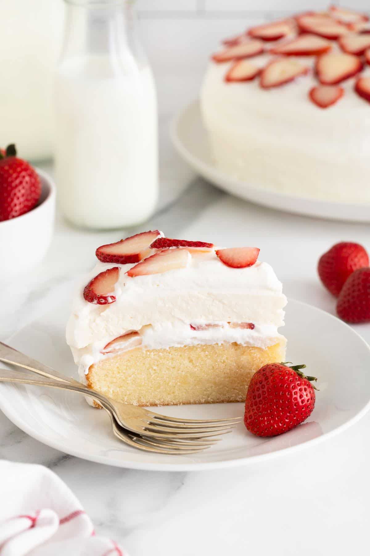 A single slice of strawberry shortcake cheesecake featuring layers of moist cake and whipped cream with fresh strawberry slices, served on a white plate with a whole strawberry and two forks, with a bottle of milk and the rest of the cake in the background