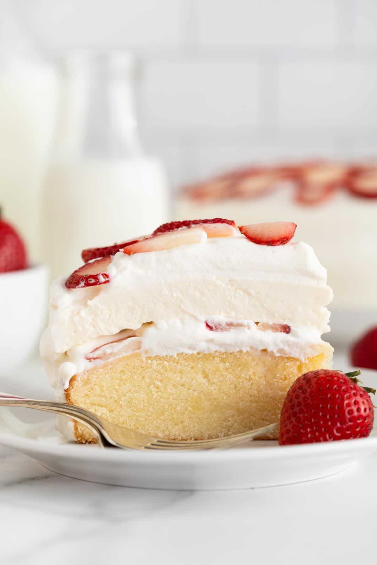 Closeup of a single slice of strawberry shortcake cheesecake featuring layers of moist cake and whipped cream with fresh strawberry slices, served on a white plate with a whole strawberry and a fork