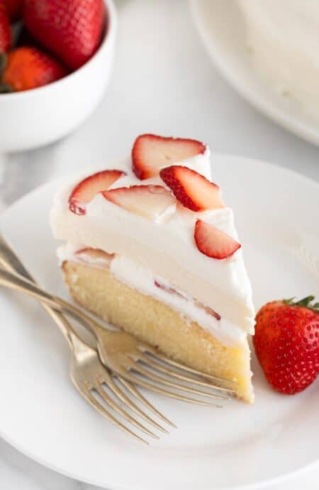 A slice of strawberry shortcake cheesecake featuring layers of moist cake and whipped cream with fresh strawberry slices, served on a white plate with a whole strawberry and two forks, with a bowl of strawberries in the background