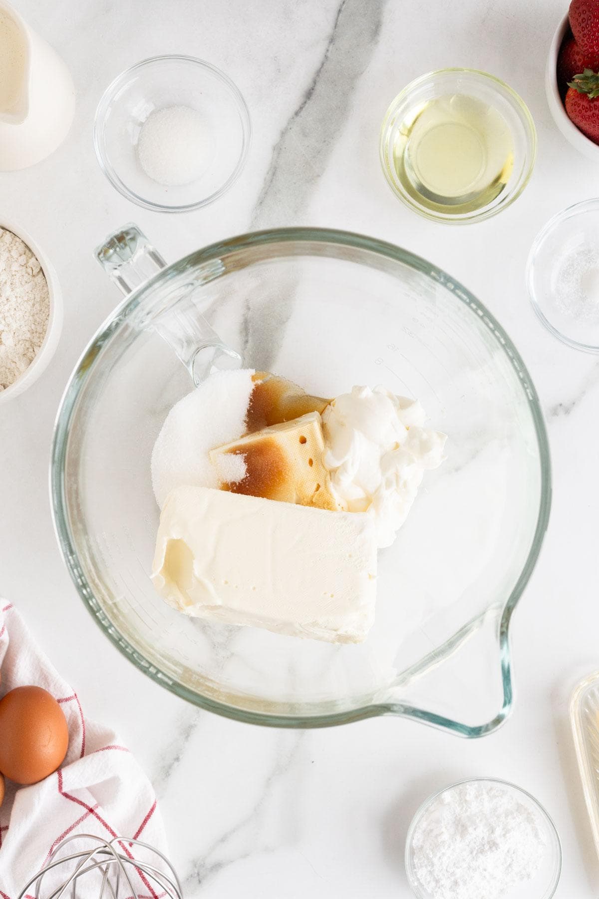 Ingredients including cream cheese, sugar, and vanilla in a glass mixing bowl, with additional baking items like eggs and flour placed around the workspace