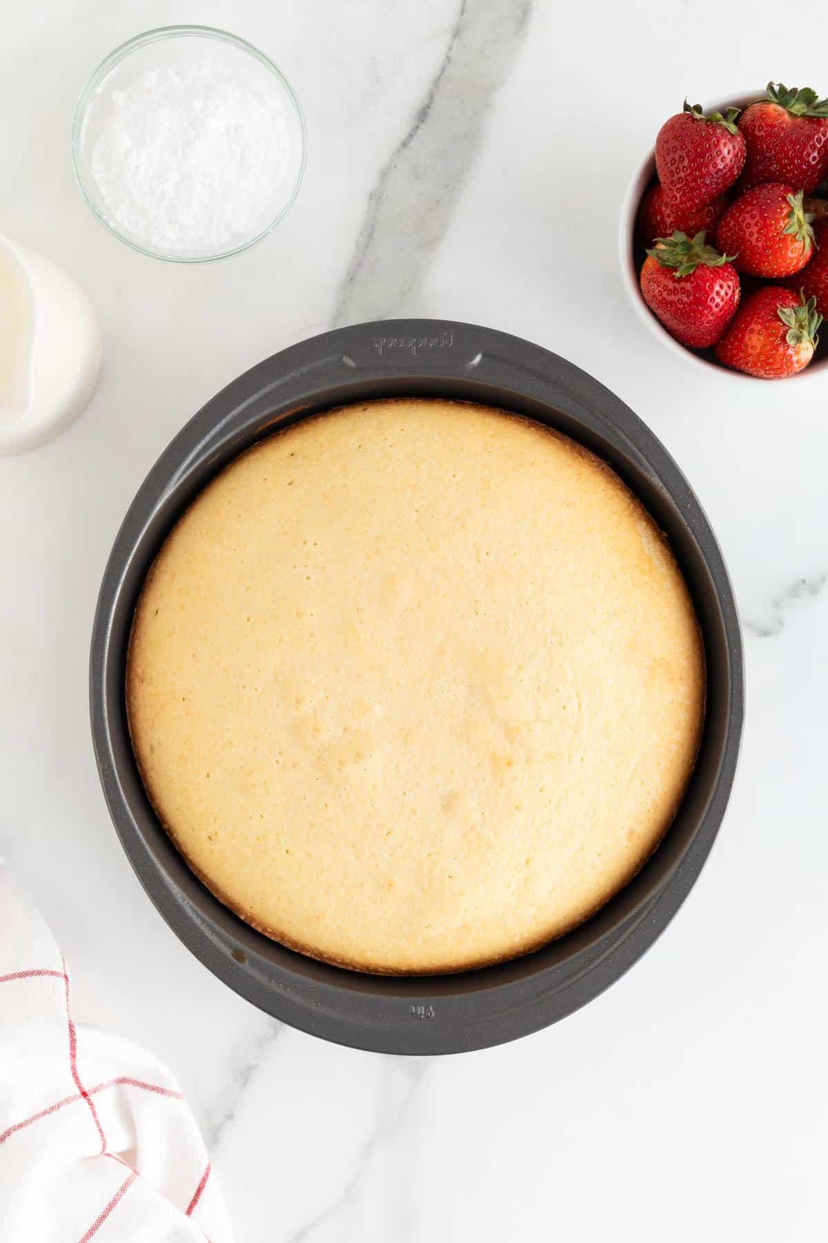 A golden-baked round cake in the pan, next to strawberries and baking ingredients