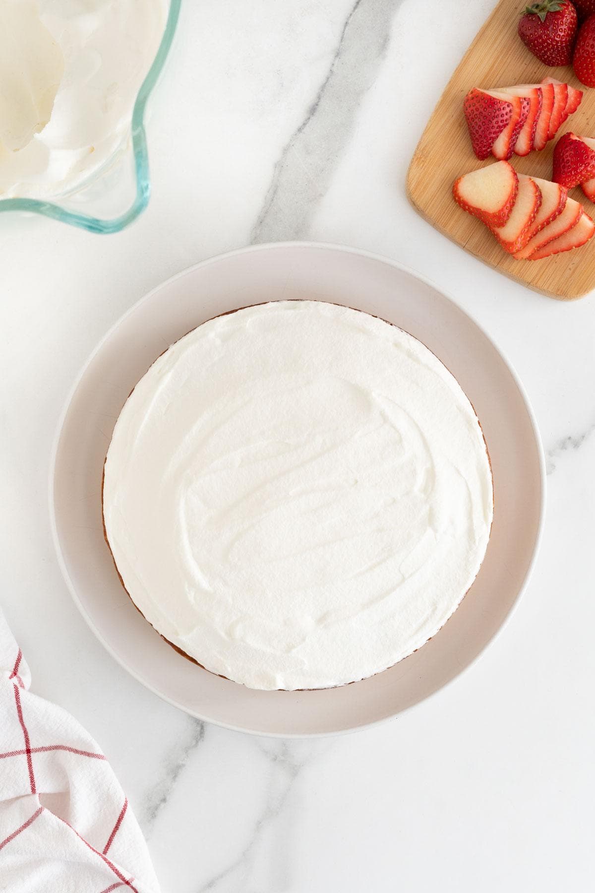 A cake layer covered with whipped cream on a plate, with sliced strawberries on a cutting board nearby