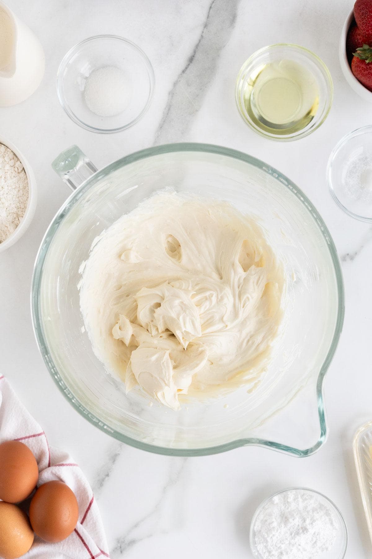 Cheesecake mixture whipped to a smooth consistency in the same glass mixing bowl, surrounded by baking ingredients