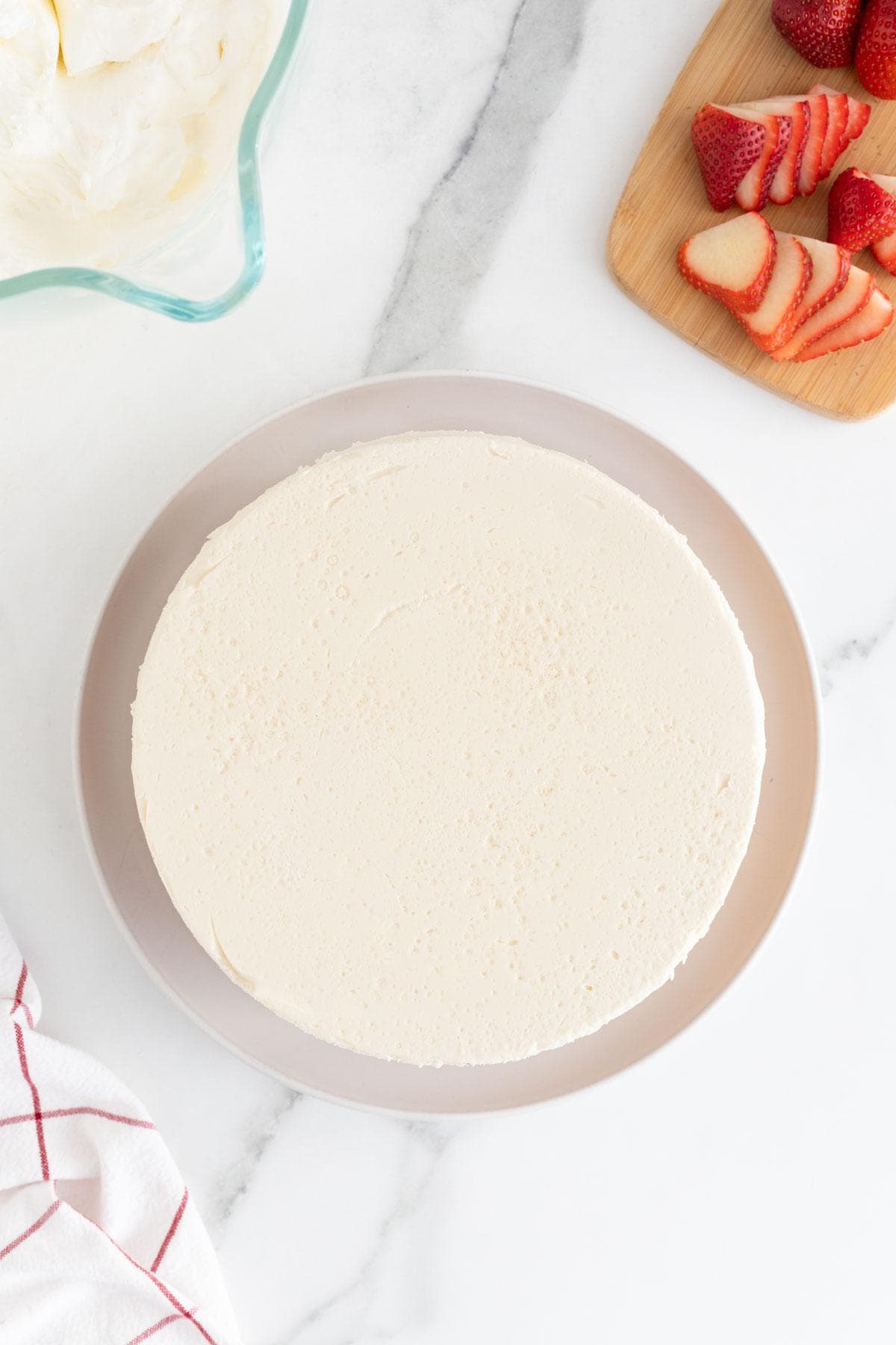 A cake layer completely covered with smooth whipped cream on a plate, strawberries on a cutting board in the background