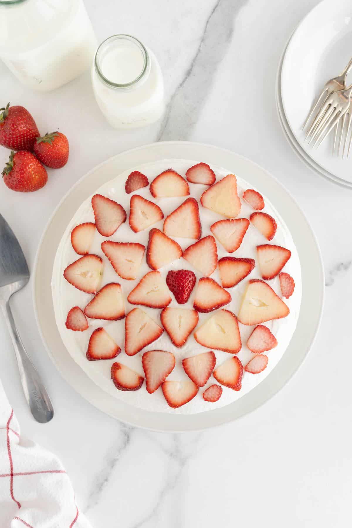 A completed strawberry shortcake cheesecake with sliced strawberries arranged decoratively on top, accompanied by milk bottles and plates with forks on the side