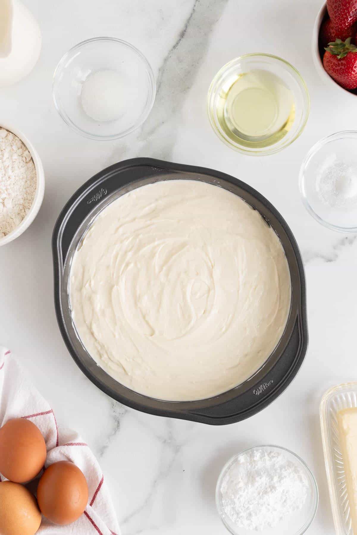 Batter evenly spread in a round cake pan, ready for baking, with eggs, oil, and other baking items visible nearby