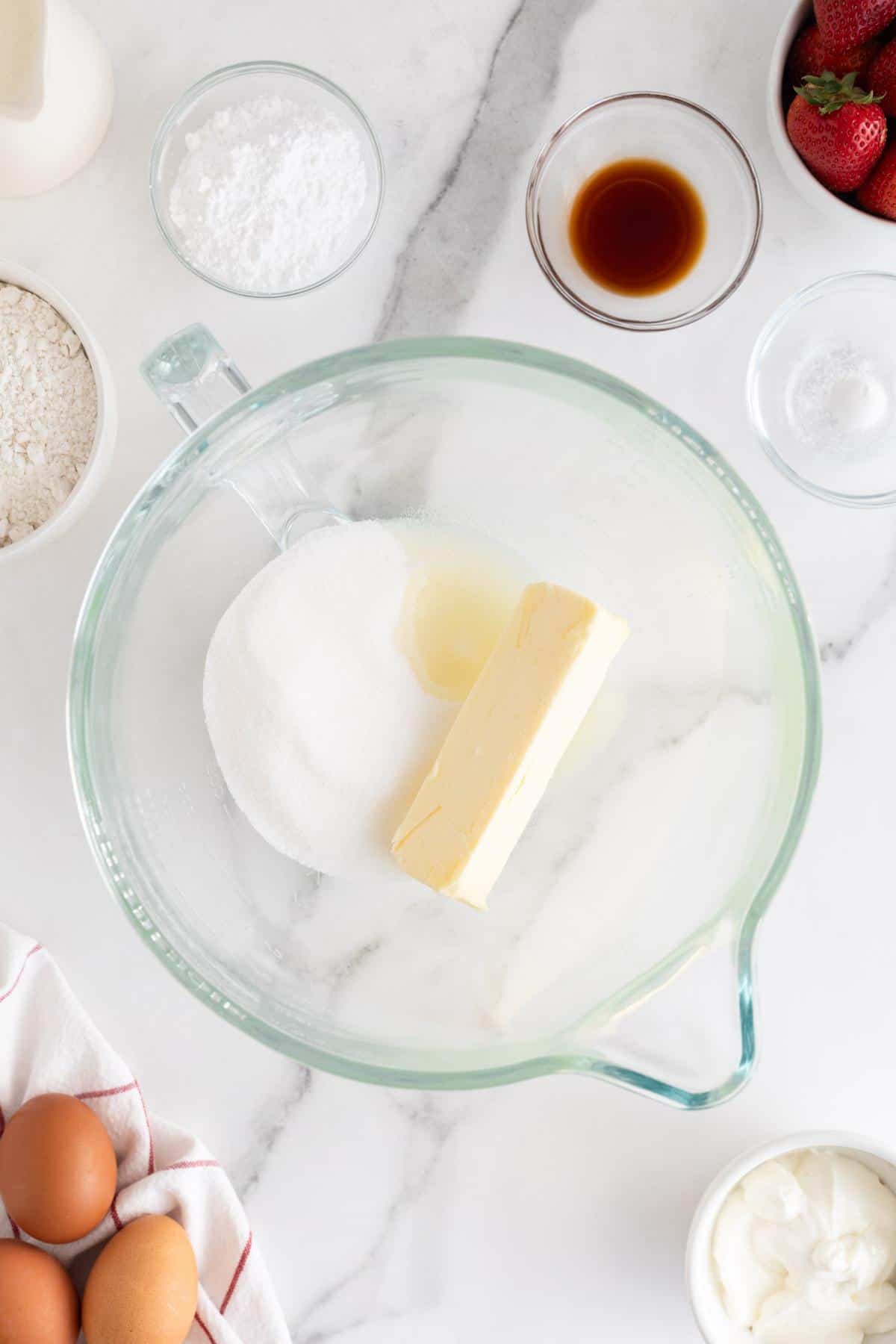 Butter and sugar in a glass mixing bowl with surrounding ingredients like eggs, strawberries, and flour arranged neatly on a white surface