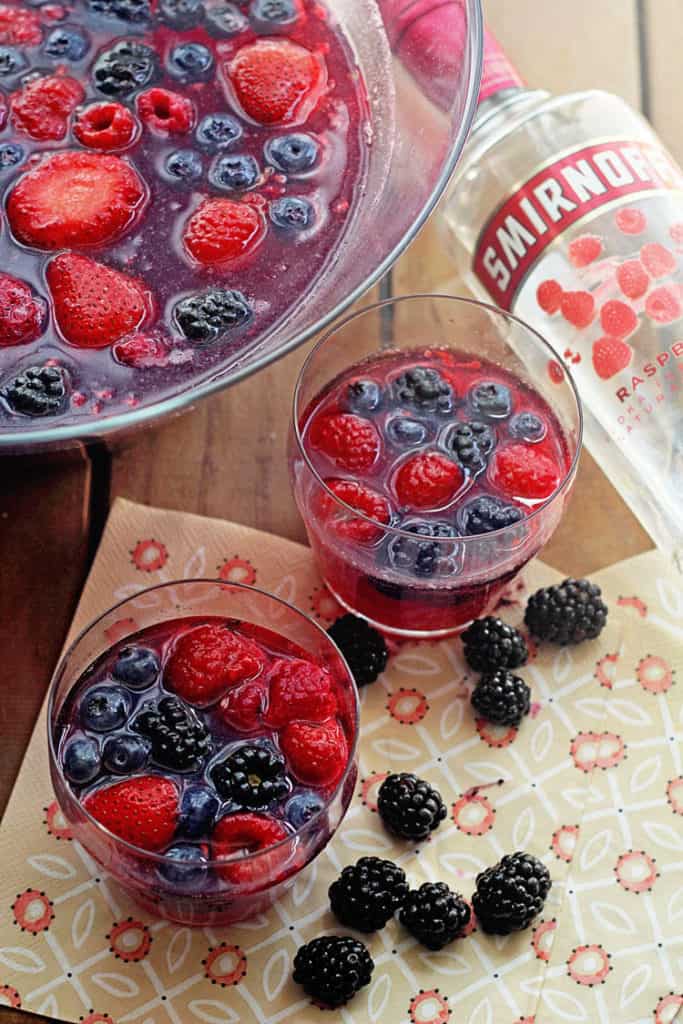 Close up of blackberry punch in a large punch bowl and two glasses of the beverage with fresh blackberries and a bottle of vodka next to it