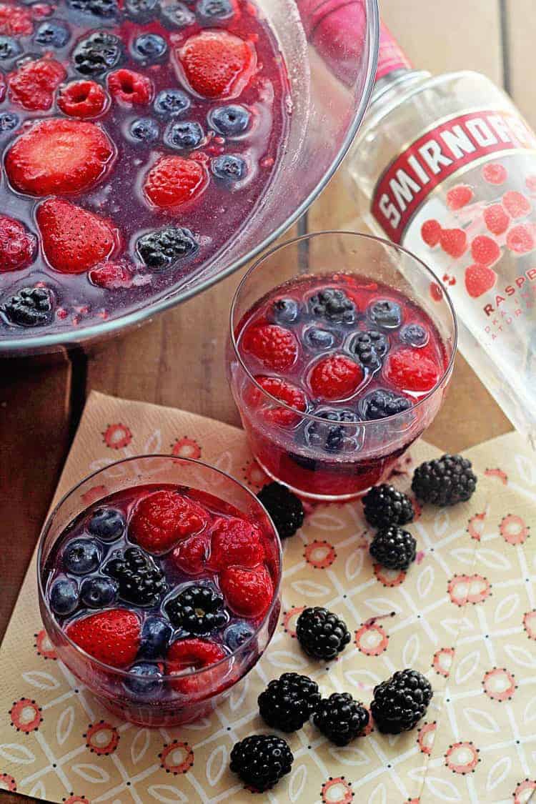 Close up of berry punch in a large punch bowl and two glasses of the beverage with fresh blackberries and a bottle of vodka next to it