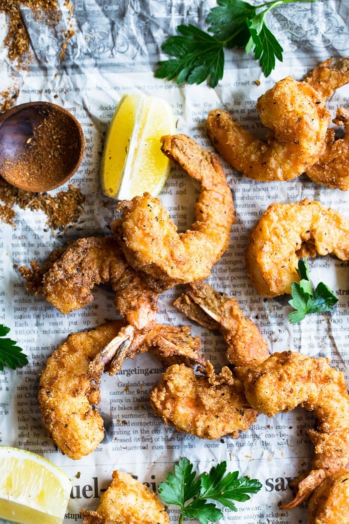 Close up of an overhead shot of Fried shrimp seasoned in cornmeal and fried to a golden brown with lemon slices and seasoning on top of newspaper