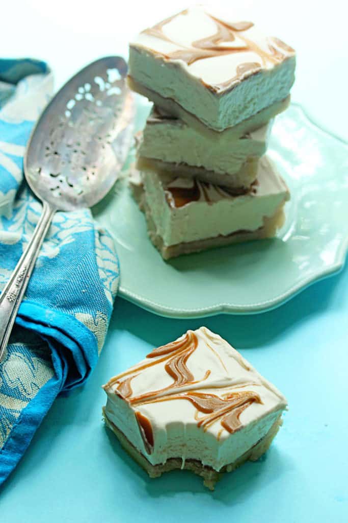 One cookie cheesecake swirl bar with a bite taken out of it and a stack of bars served on a white plate in the background