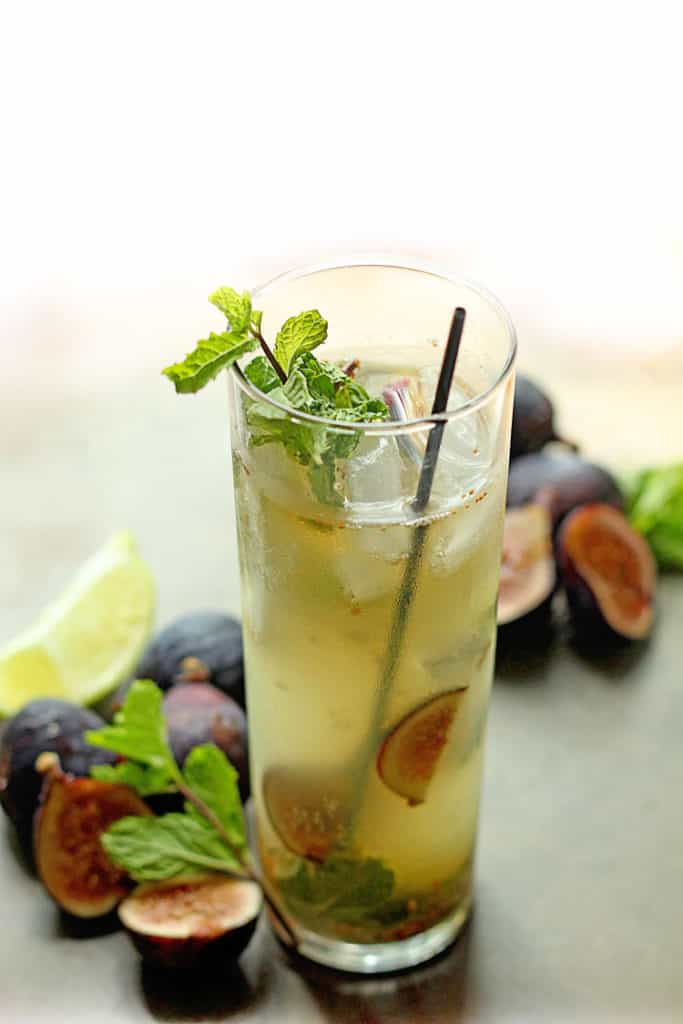 Close up of a fig mojito with a skinny, black straw and mint in it and sliced figs and lemon around it
