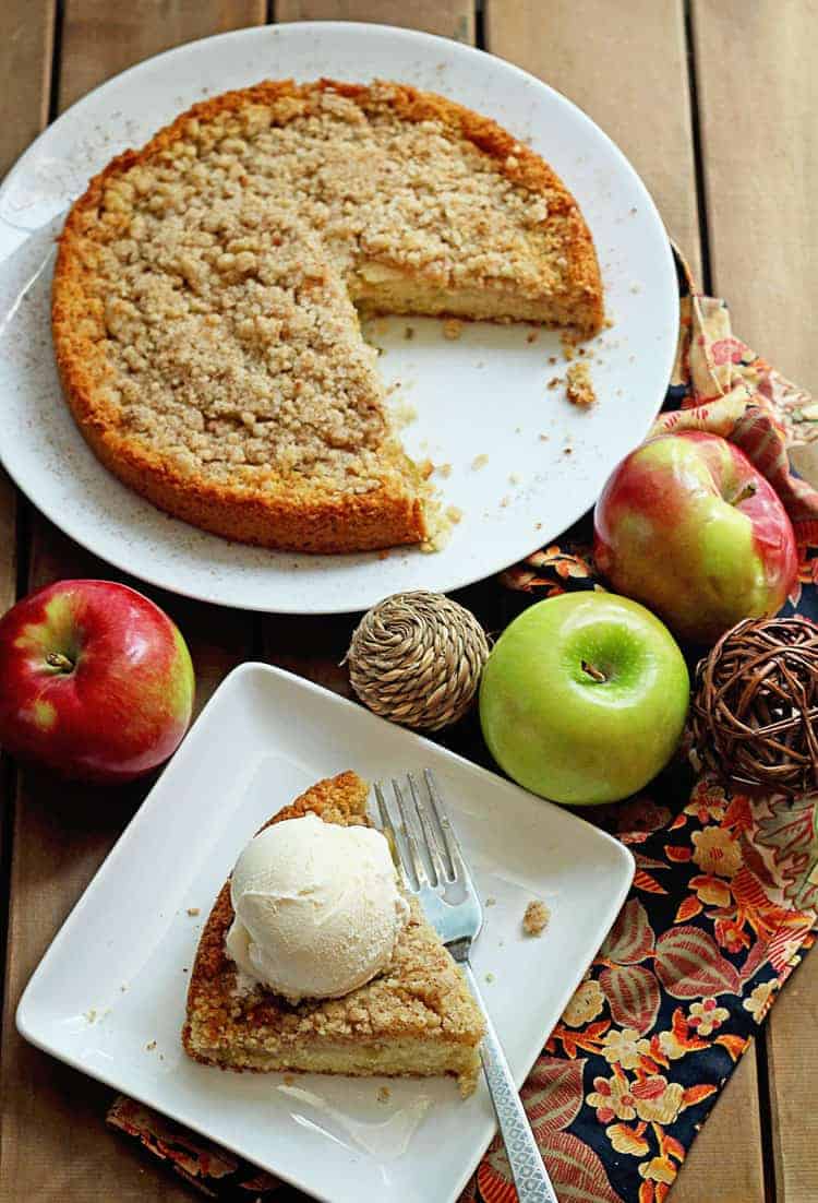 An apple cake served on a circular, white plate with a piece missing and a slice of the cake served on a square, white plate with a fork and topped with a scoop of vanilla ice cream next to it along with three whole apples as decoration