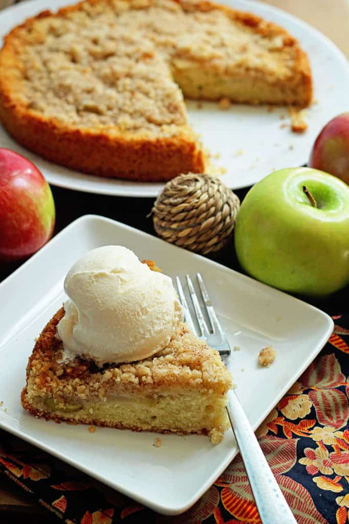 Close up of maple apple cake served on a circular, white plate with a piece missing and a slice of the cake served on a square, white plate with a fork and topped with a scoop of vanilla ice cream next to it along with three whole apples as decoration