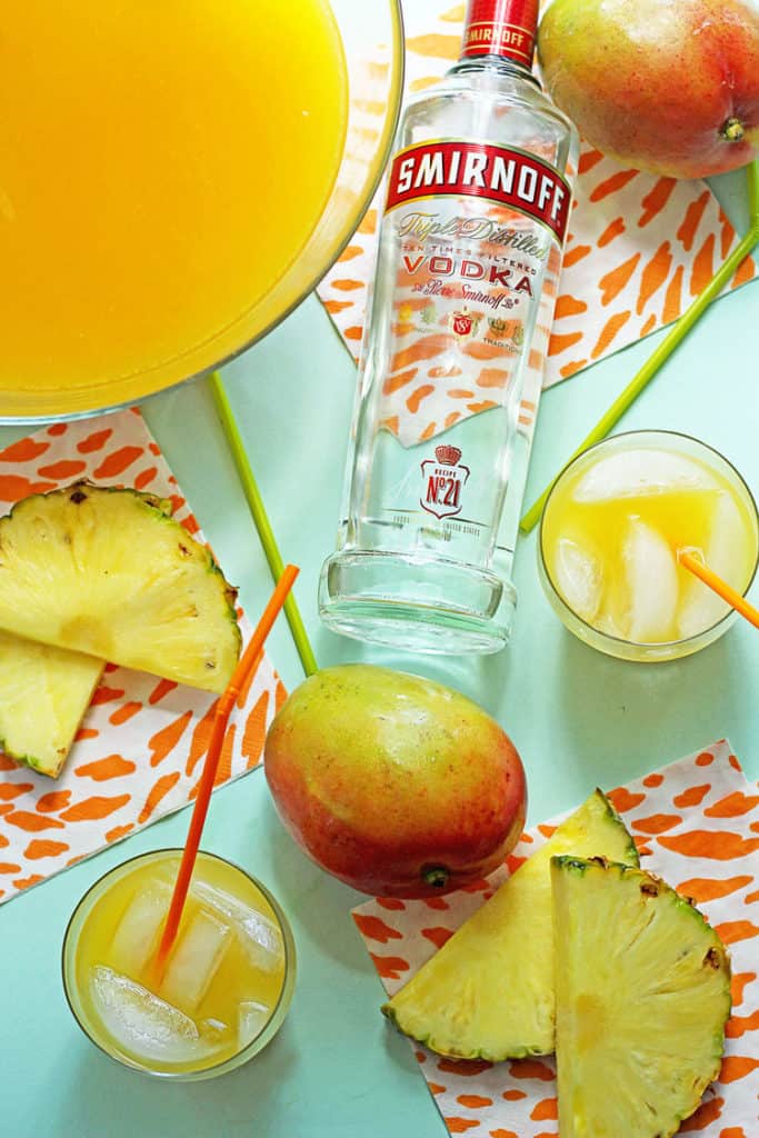 Overhead shot of tropical pineapple punch contained in a glass punch bowl and two glasses with straws with pineapple slices, a whole mango and a bottle of vodka decorating the scene
