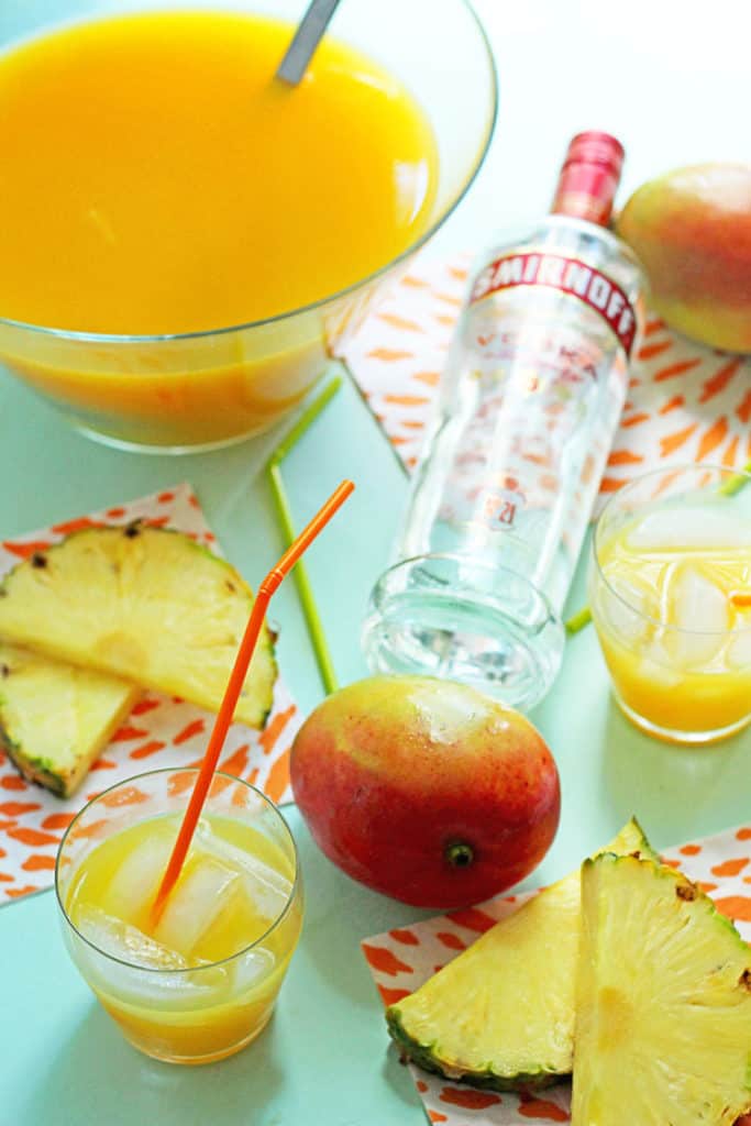 Tropical Pineapple Punch contained in a glass punch bowl and two glasses with straws with pineapple slices, a whole mango and a bottle of vodka decorating the scene