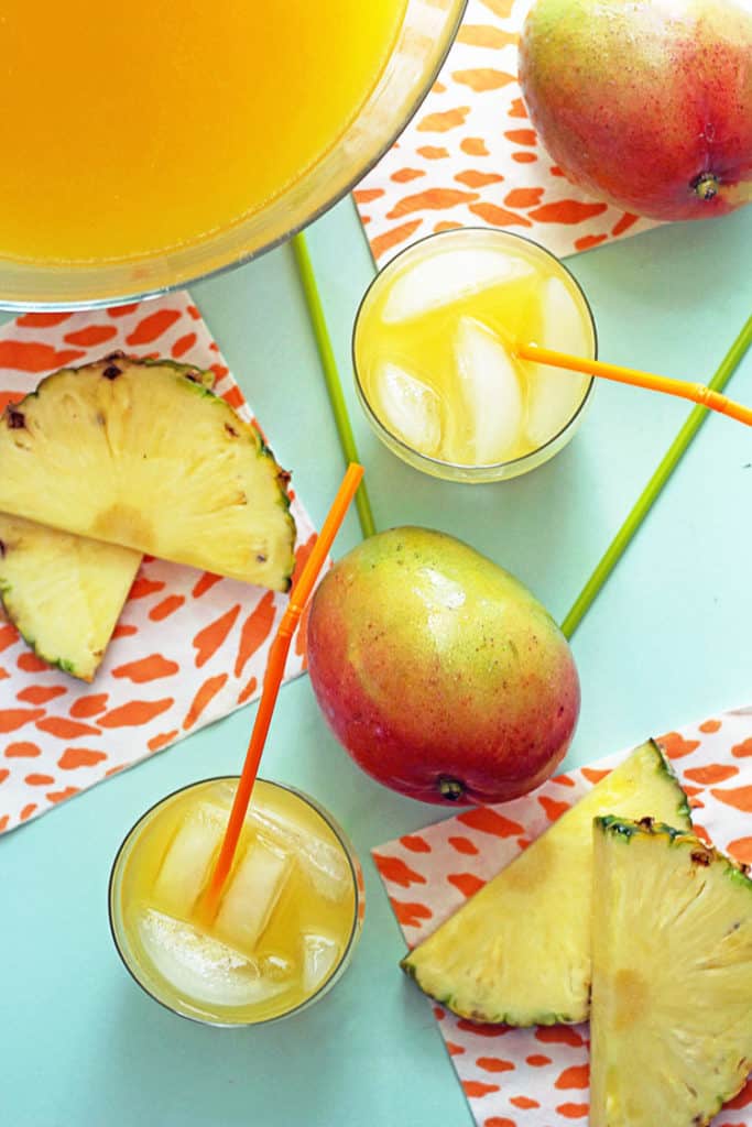Overhead shot of tropical pineapple punch contained in a glass punch bowl and two glasses with straws with pineapple slices and a whole mango decorating the scene