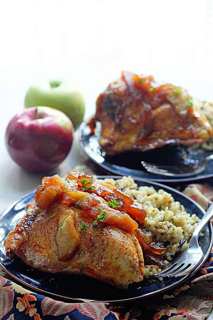Apple Cider Glazed Chicken served on a black plate with rice and a fork and a couple of fresh apples another plate of the dish in the background