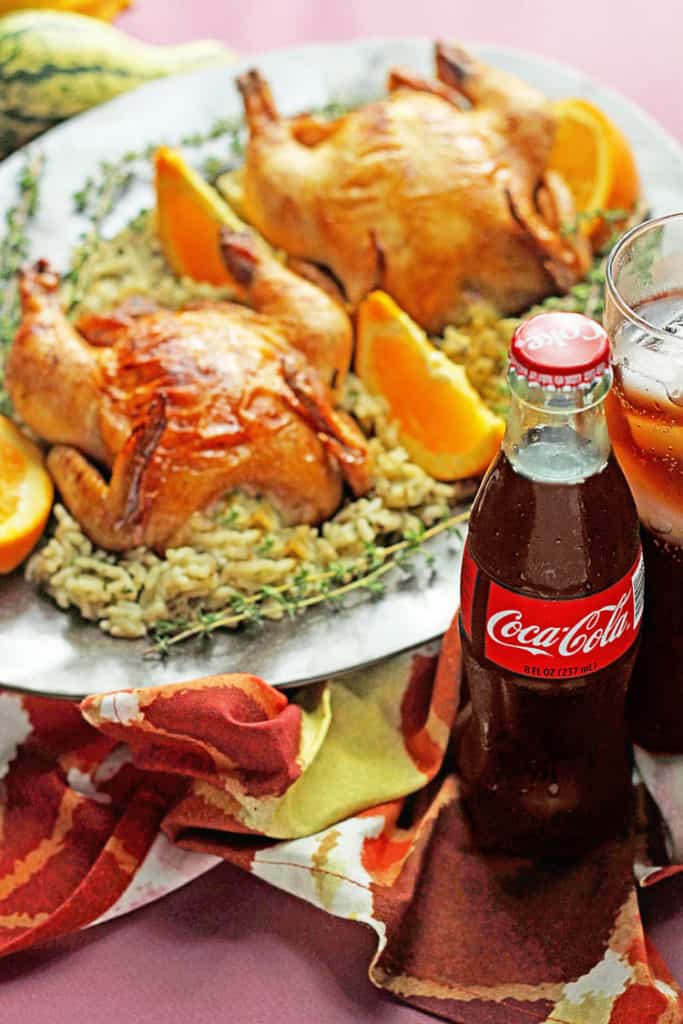 Close up of a bottle of coca-cola and glass full of the beverage with two Coca-Cola Glazed Cornish Hens in the background