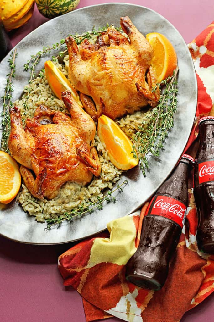 Two Coca-Cola Glazed Cornish Hens served on a silver platter with rice and orange wedges and two coke bottles next to it