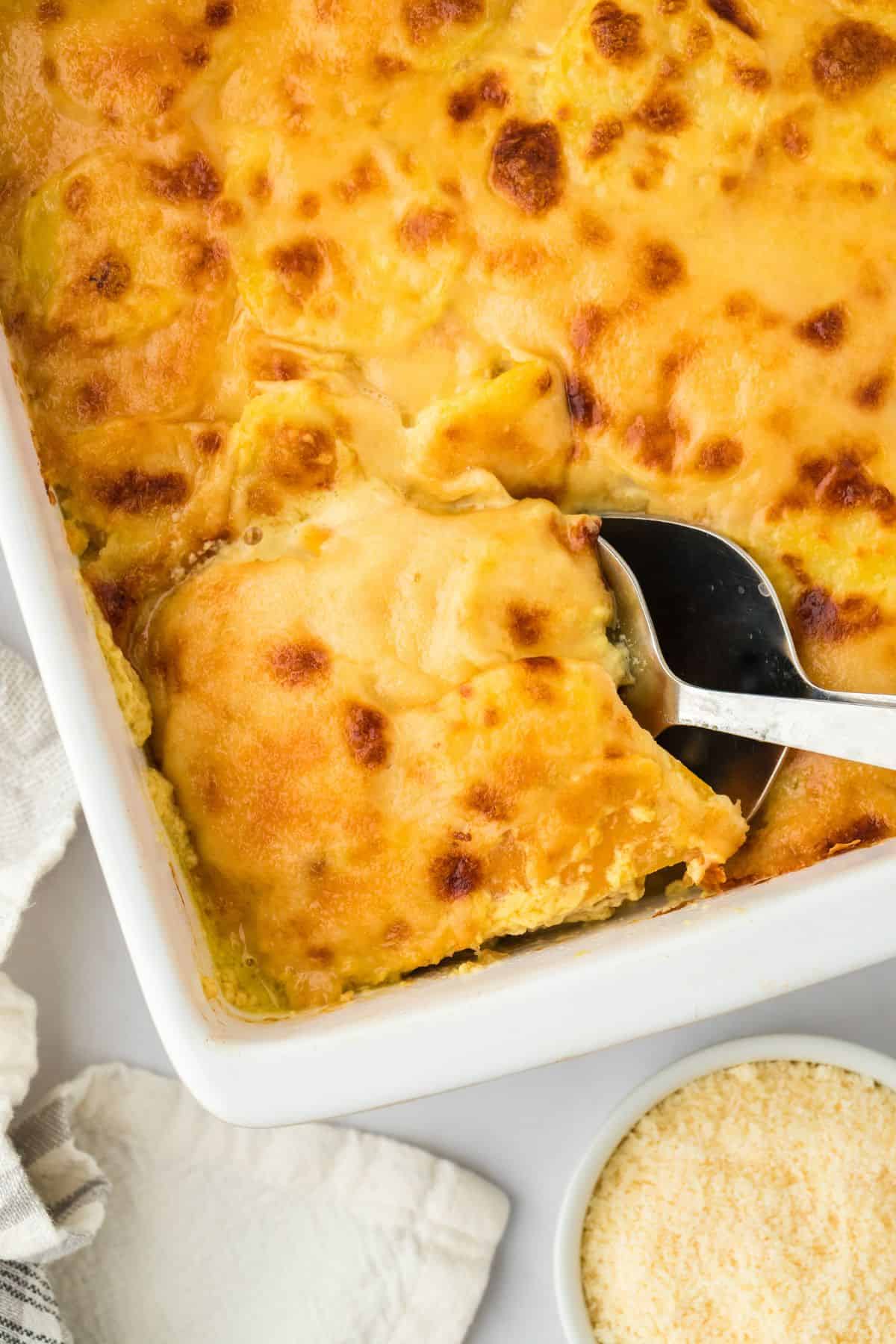 Closeup of baked potato and squash gratin in a white baking dish, with a spoon lifting a portion and a bowl of parmesan cheese nearby