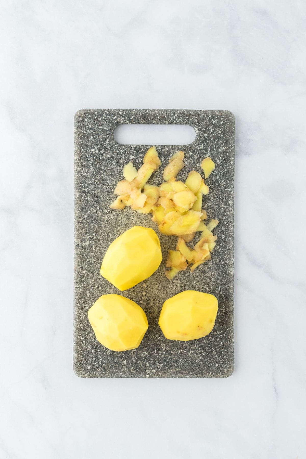 Peeled potatoes on a grey cutting board with potato skins set aside