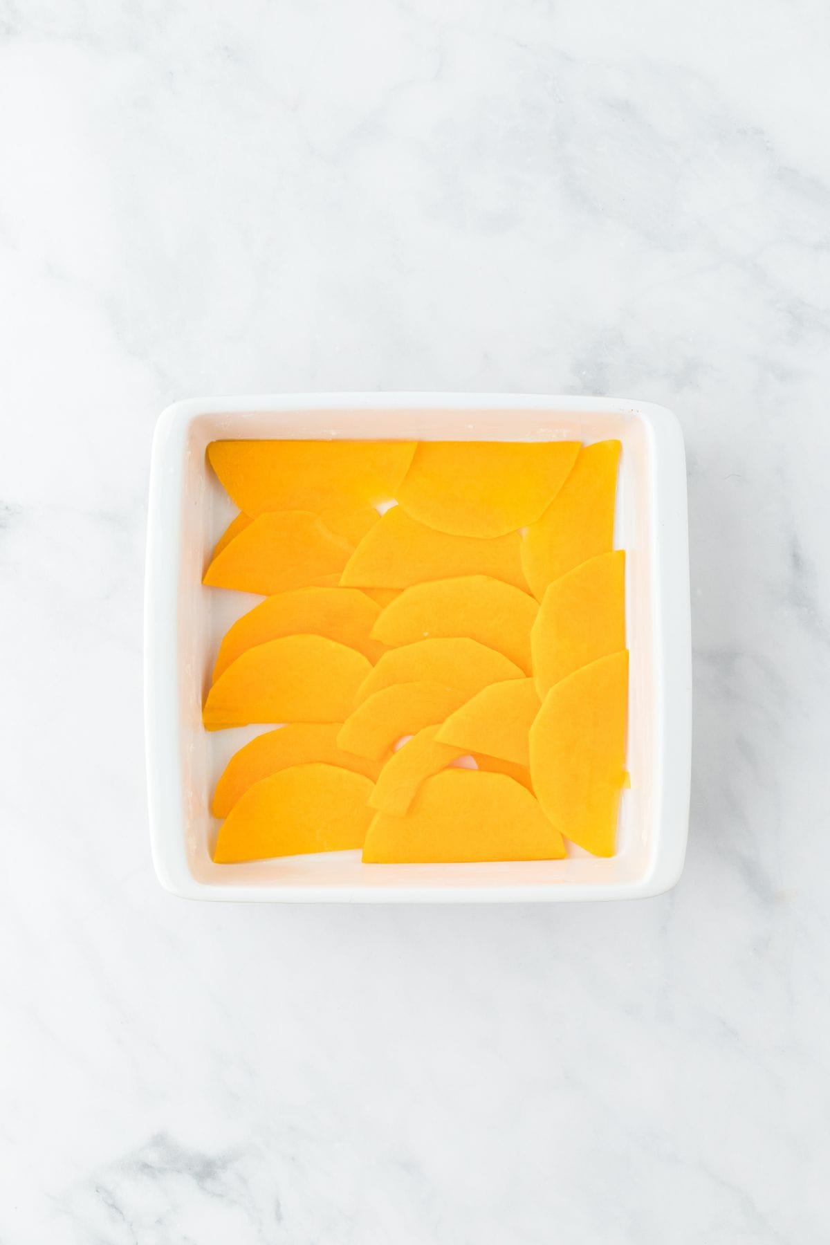 A white baking dish with a single layer of thinly sliced butternut squash