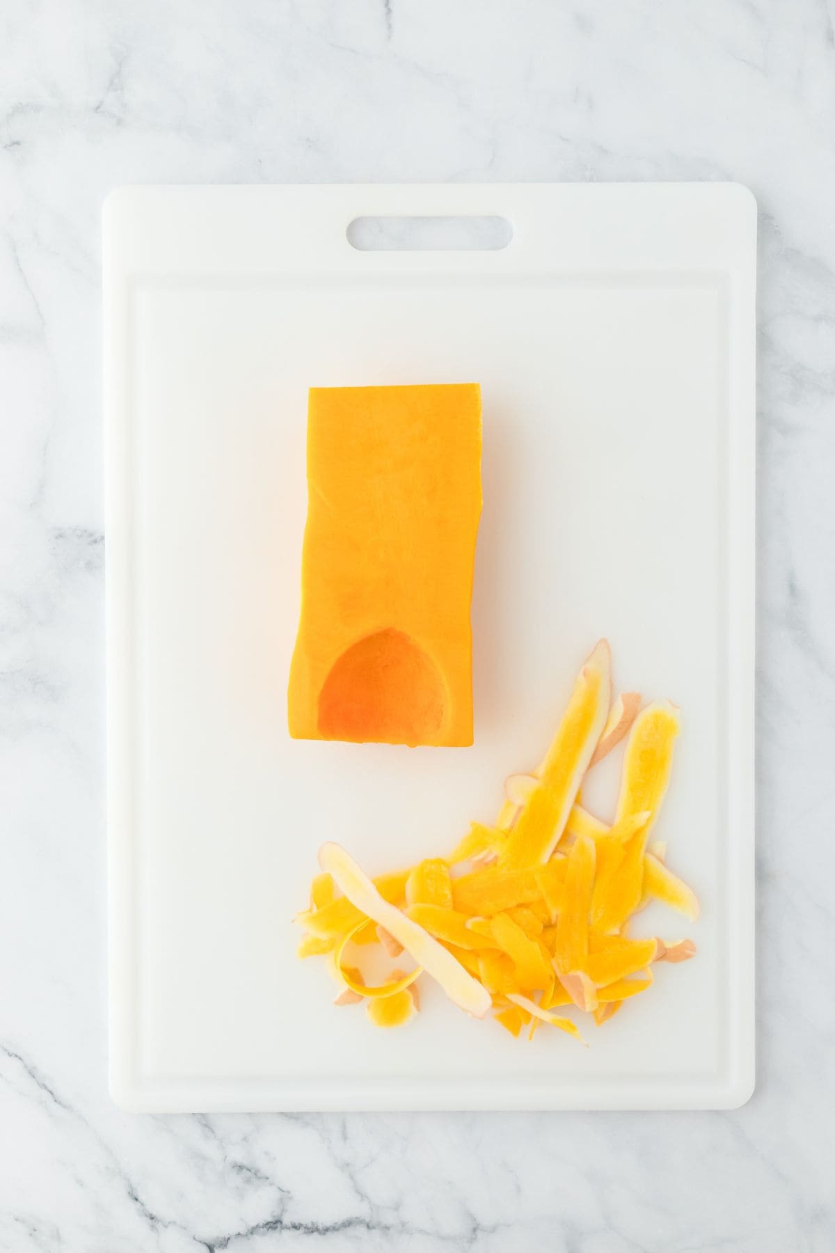 A peeled butternut squash on a white cutting board with its peels next to it