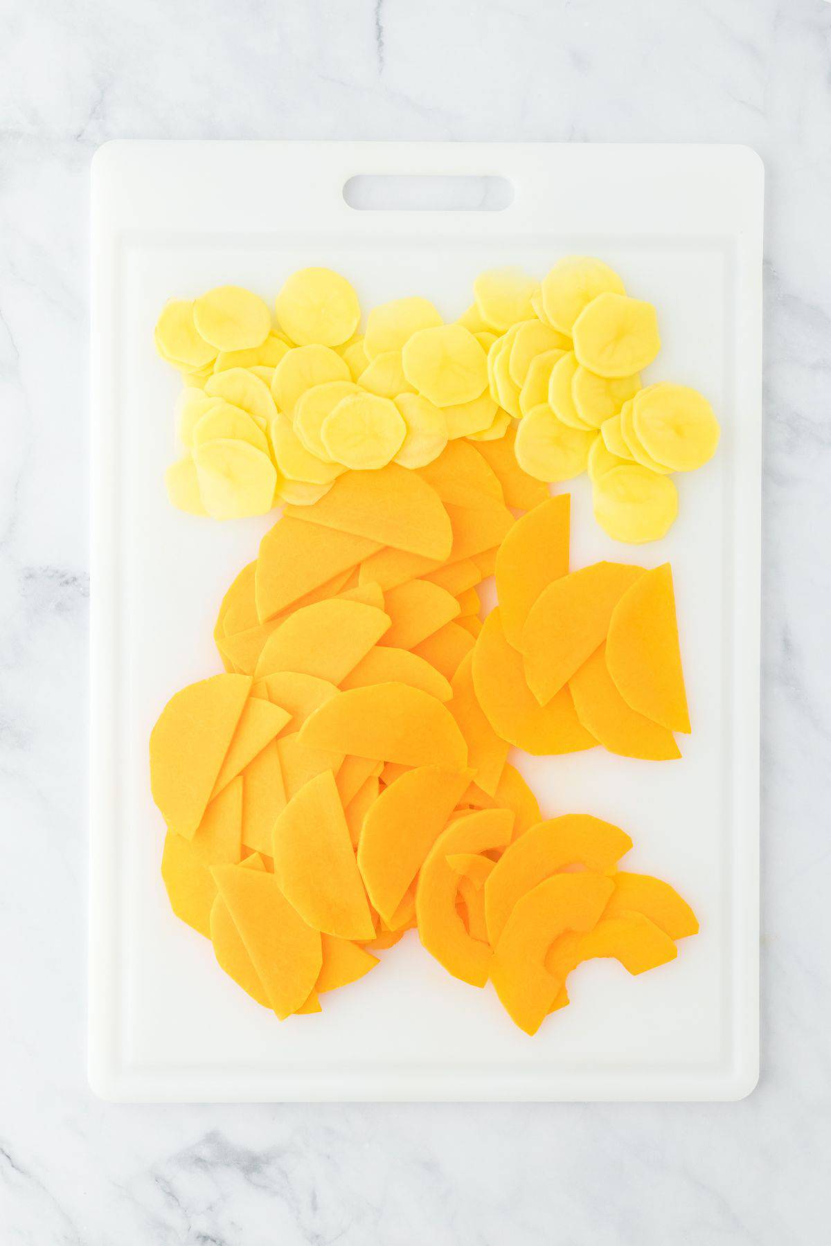 Thinly sliced yellow potatoes and orange squash arranged on a white cutting board