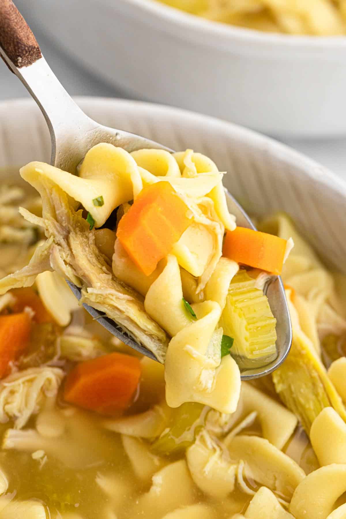 A spoon holding a spoonful of slow cooker chicken noodle soup up over the a bowl of more soup.