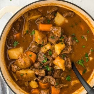 A simple beef stew recipe in a pot with a large spoon stirring on a white background with parsley in the background