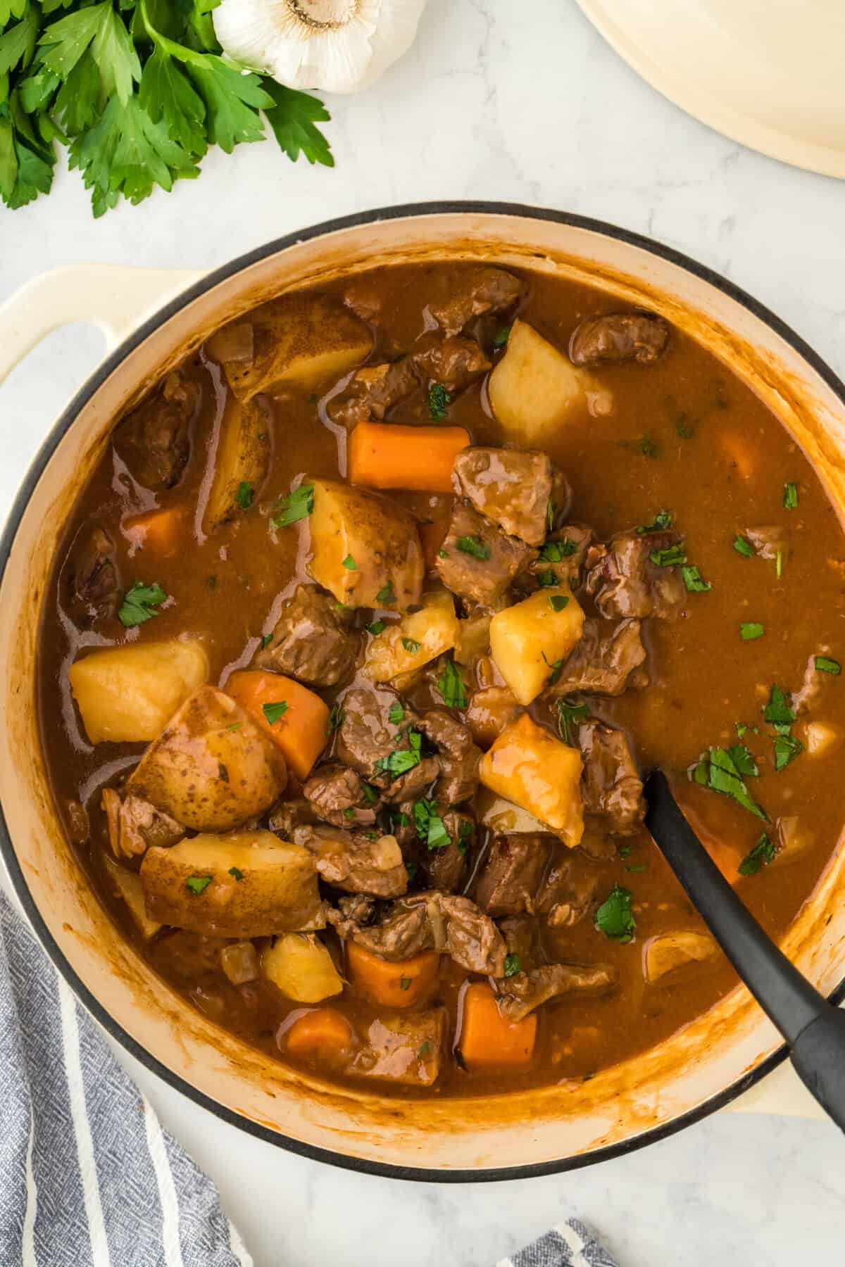 A simple beef stew recipe in a pot with a large spoon stirring on a white background with parsley in the background