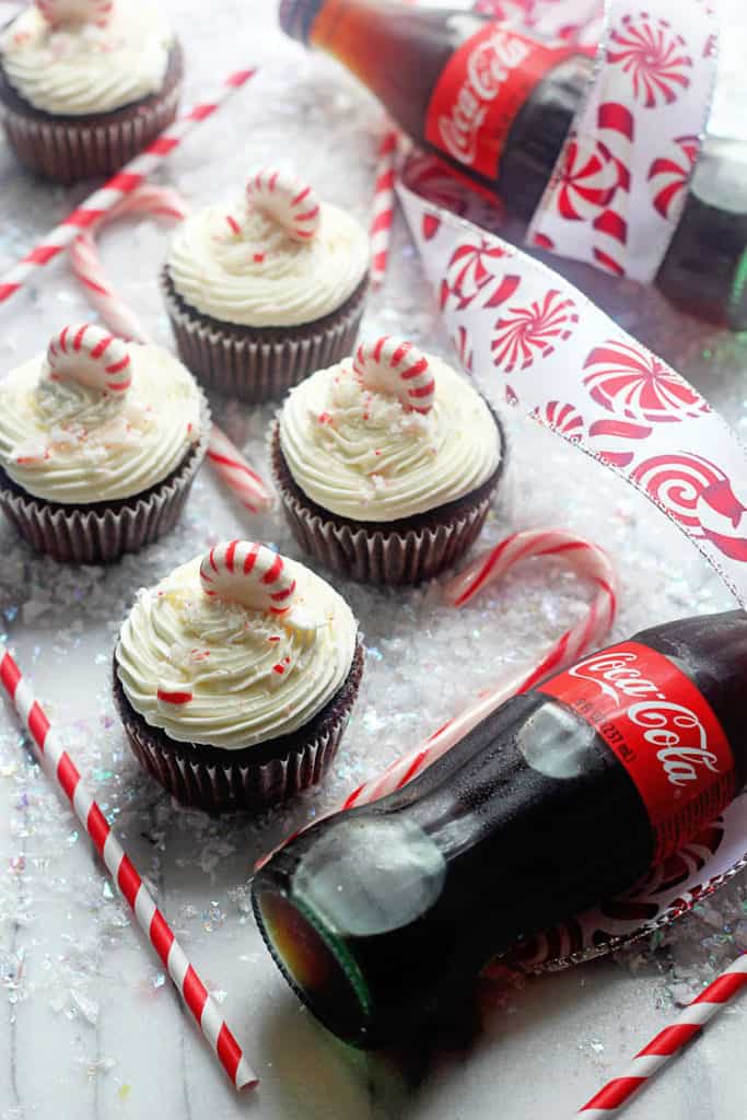 Coca Cola Chocolate Cupcakes with Peppermint Buttercream topped with peppermint candy surrounded by a couple bottles of Coca-cola soda and candy canes. 
