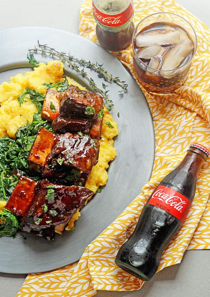 Three braised beef ribs on a bed of spinach and polenta next to cola cola bottle ready to serve for dinner