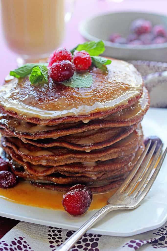 Gingerbread Pancakes recipe drenched in eggnog syrup topped with mint and fresh cranberries on a white plate with a fork next to it.