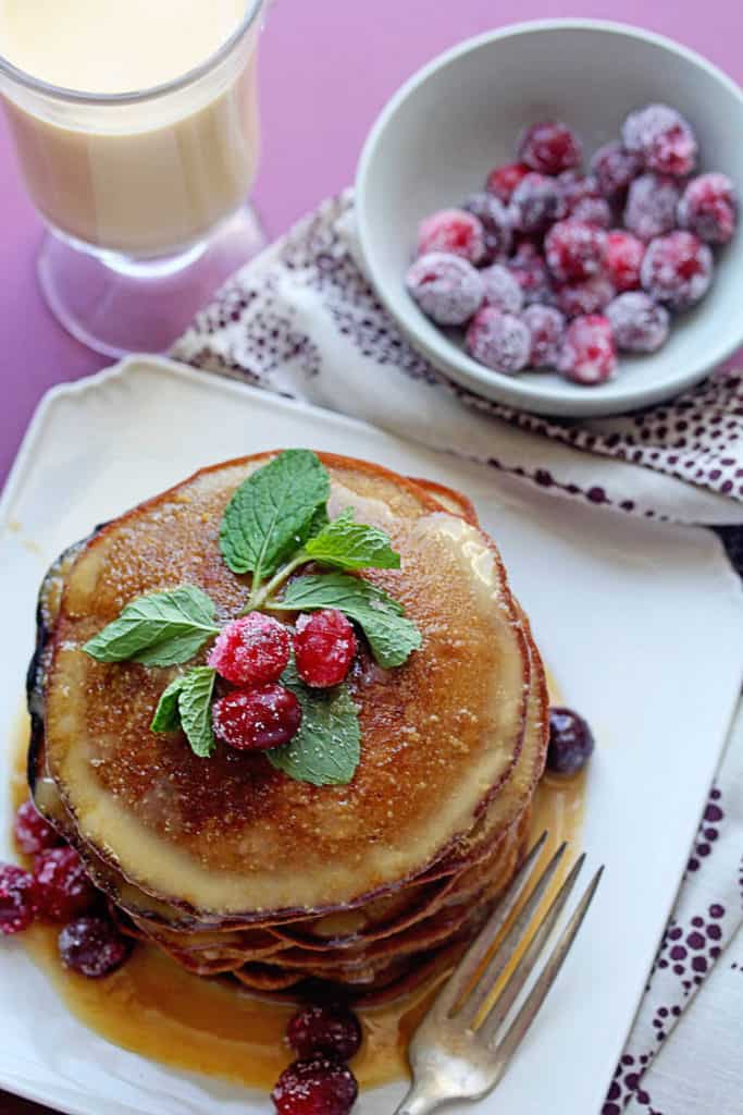 Overhead shot of gingerbread pancakes topped with eggnog syrup, fresh mint and cranberries sitting on a white plate with a fork next to it.