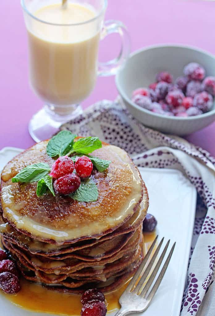 Gingerbread Pancakes with Eggnog Syrup topped with cranberries and mint as a garish sitting on a white plate next to a cup of eggnog and a bow of cranberries.