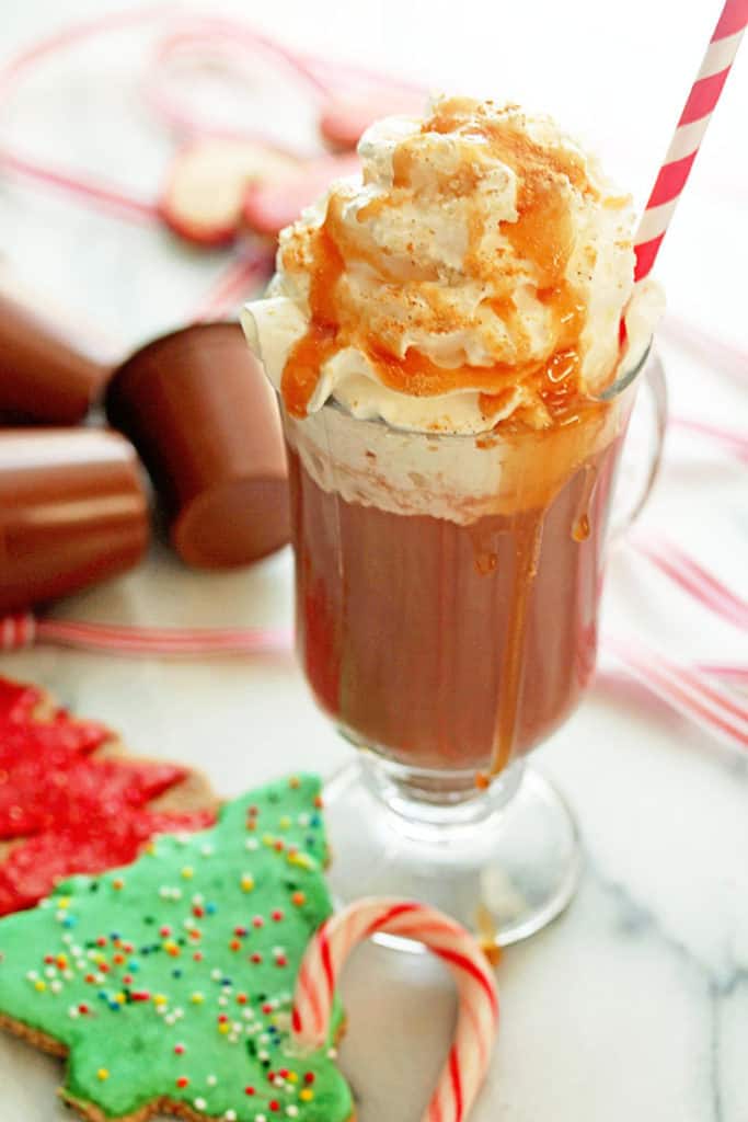 Close up of Salted Caramel Ginger Hot Cocoa served in a glass mug with a white and red striped straw in it and Christmas cookies and a candy cane next to it