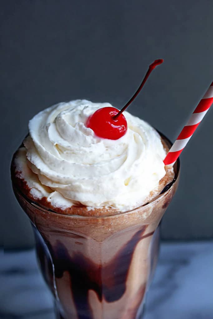 Close up of a skinny chocolate milkshake topped with whipped cream and a cherry with a white and red striped straw in it