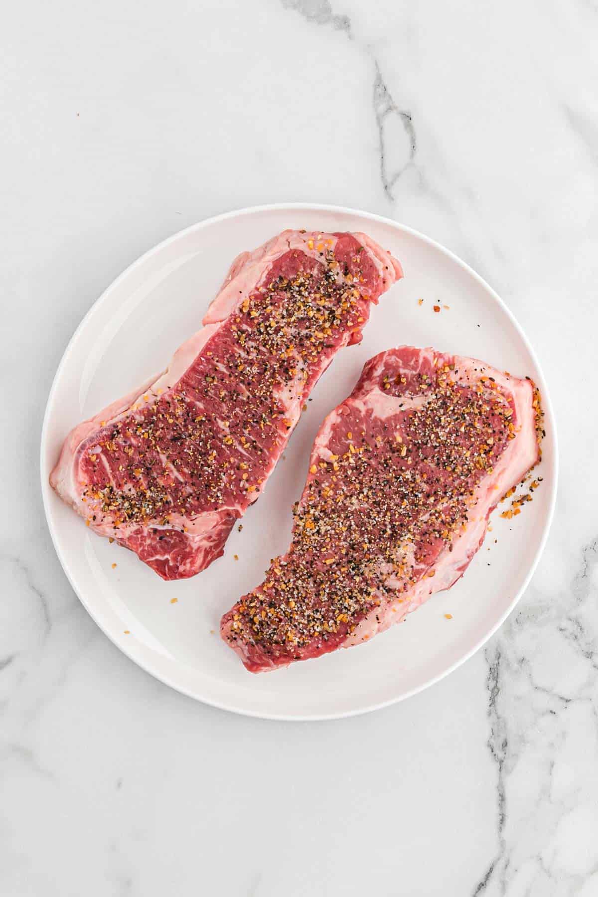 Steaks on a plate are sprinkled with seasoning before cooking.