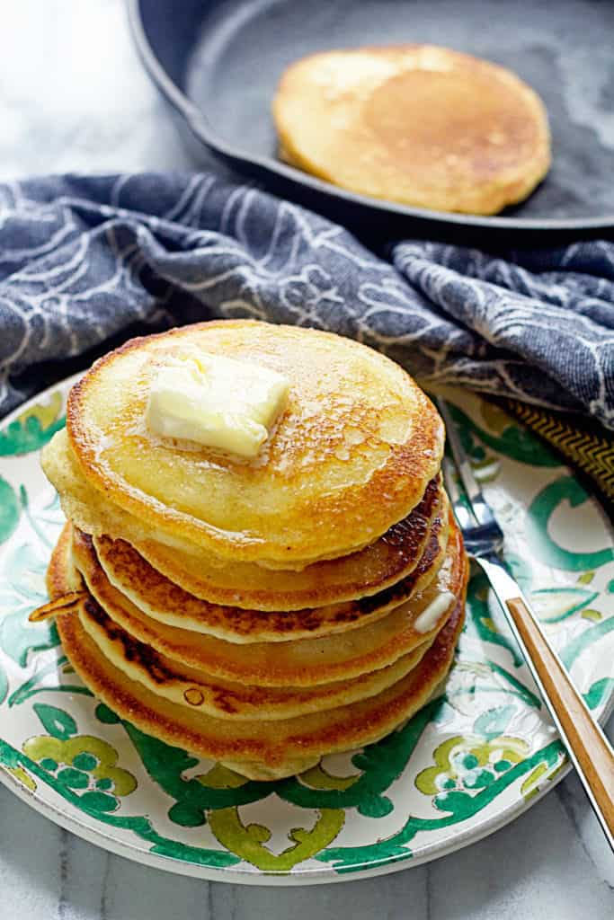 A stack of Hoe Cakes topped with butter and sitting on a green and white plate with a fork and more in the background in a skillet