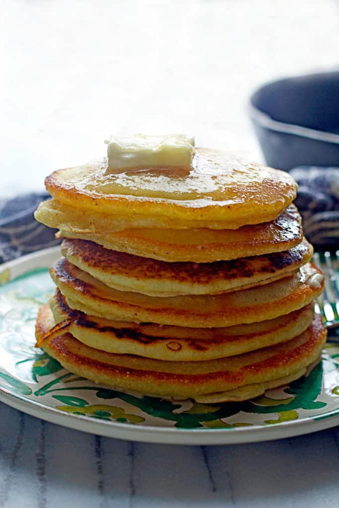 A stack of Johnny Cakes topped with butter and sitting on a green and white plate with a fork